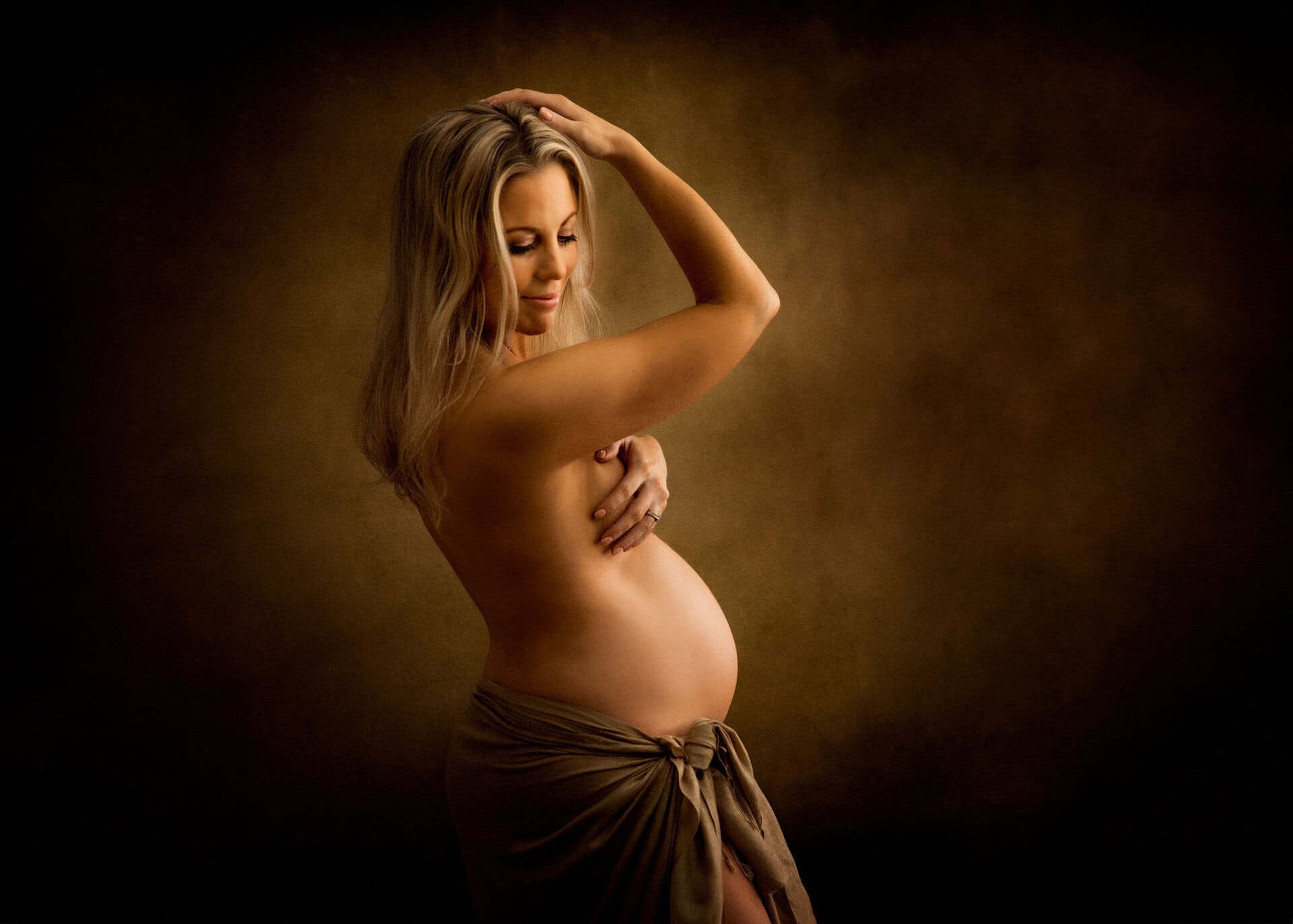 Fine art maternity photographers in Crawley. Beautiful pregnant lady poses against a dark background, embodying the elegance of Sussex maternity portraits. She modestly covers her chest with one arm while a draped cloth gracefully encircles her waist.