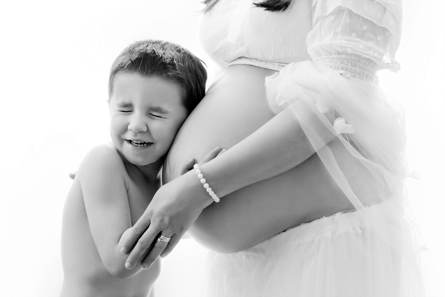 Award-winning Crawley maternity photographers. A young child smiles with eyes closed, hugging a pregnant womans belly in this tender Sussex maternity portrait. Both are dressed in light, airy clothing against a pristine white background, capturing a moment of pure joy and anticipation.
