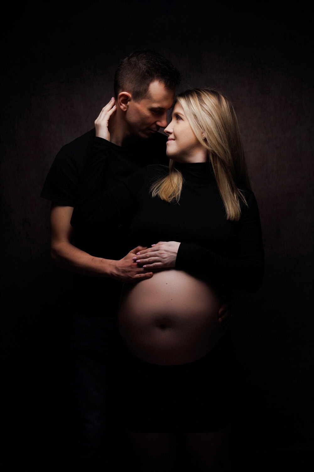 Award winning maternity photographers in Crawley. A couple poses lovingly in front of a dark background. The woman, pregnant, holds her partners hand on her belly as they share an intimate glance. Their black outfits enhance the timeless elegance commonly seen in Sussex maternity portraits.