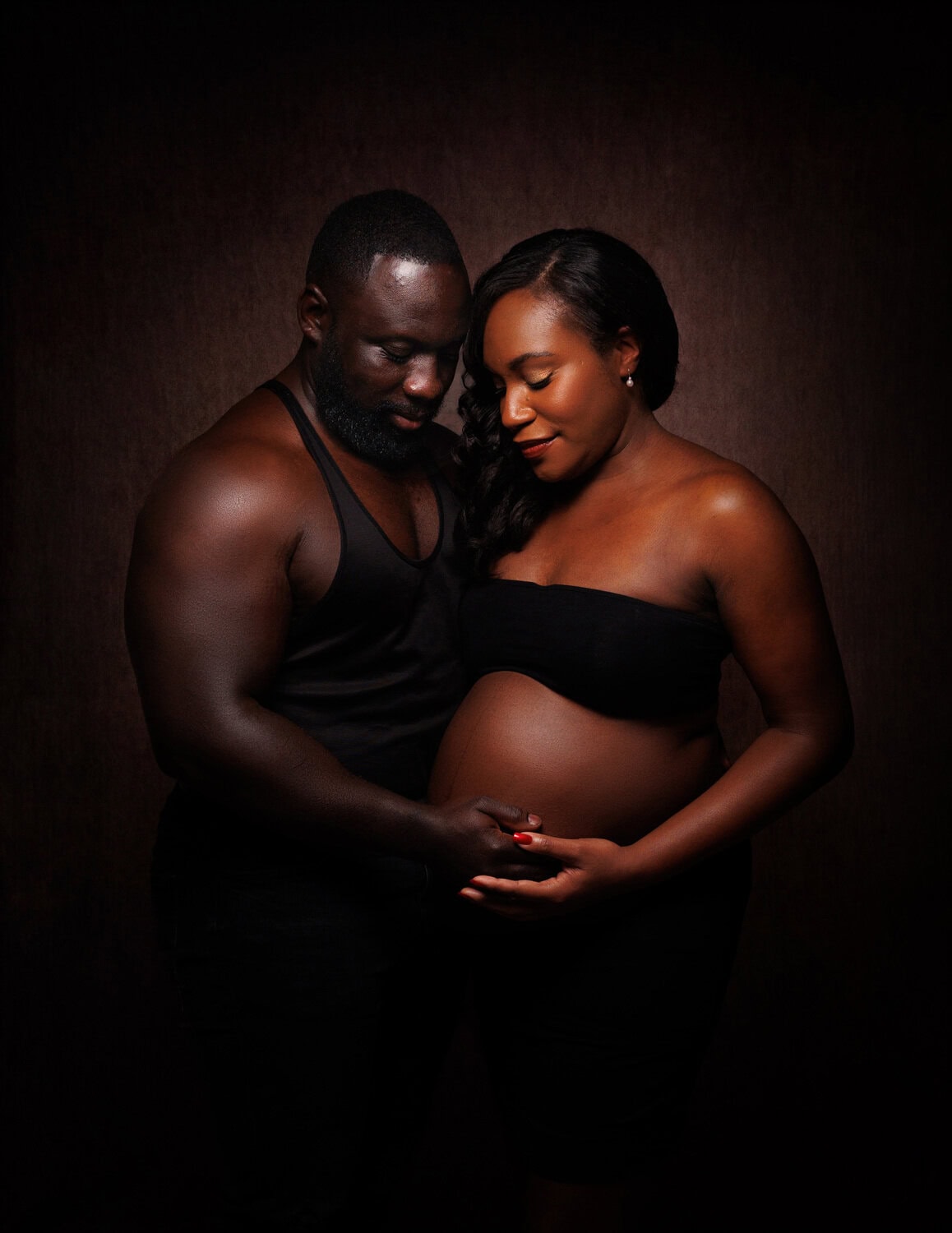 A couple stands together against a dark background, the mans hands gently resting on his partners pregnant belly. Both dressed in black, they embody the elegance of Sussex maternity portraits.