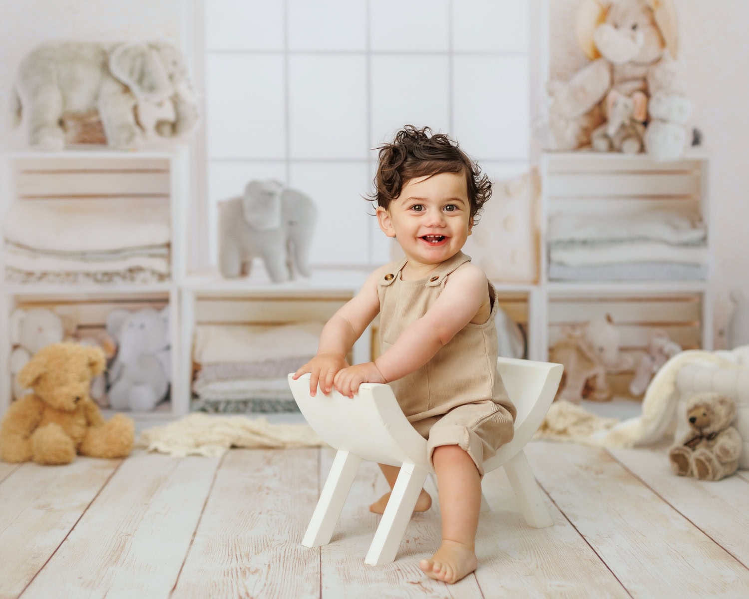 In a serene nursery setting, a child in a beige outfit sits comfortably on a white chair, surrounded by plush stuffed animals and shelves lined with neatly folded linens. This charming scene captures the essence of Sussex baby photographers talent against a windowed backdrop.