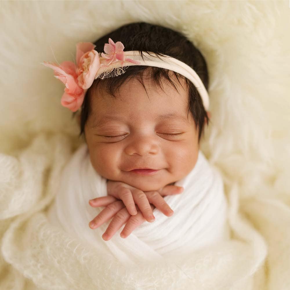 Award winning Crawley newborn photographers. A smiling baby with dark hair, safely wrapped in a white blanket, and adorned with a peach headband featuring flowers, all set against a soft, creamy background.