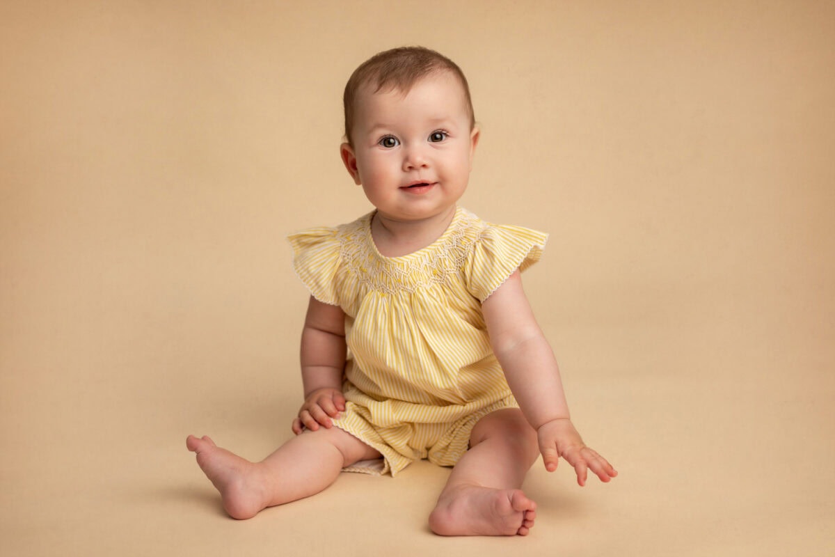 Baby sitting on a beige surface in a yellow striped outfit, gazing forward with a neutral expression. For more moments like this, contact us.