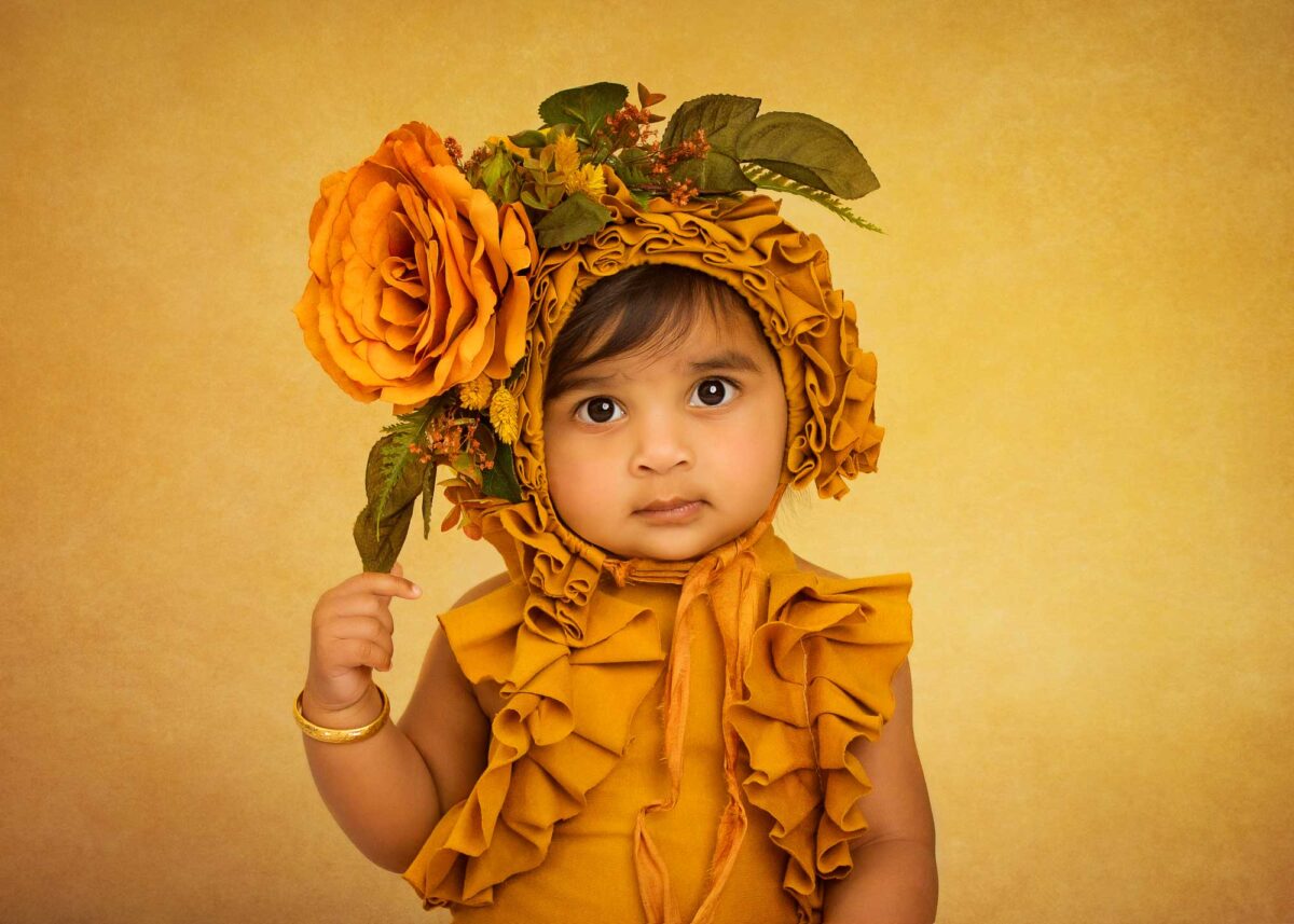 A toddler sits charmingly in a ruffled, mustard-colored outfit with a floral headdress, holding a large orange flower against a matching mustard background, capturing the essence of elegant sitter baby portraits.