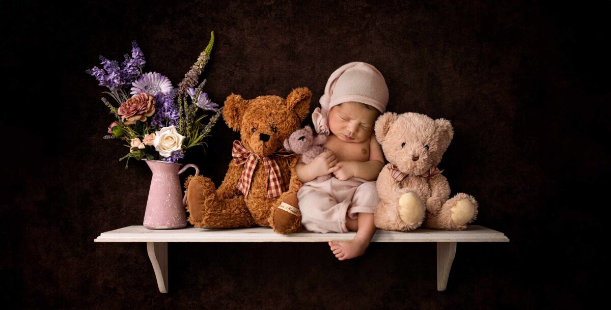 Fine Art newborn photoshoot featuring a sleeping baby in a pink outfit with a hat rests on a shelf, surrounded by three teddy bears. This image was created in complete safety as a composite image, with baby laying safely on his back on a soft beanbag, and edited in Photoshop.