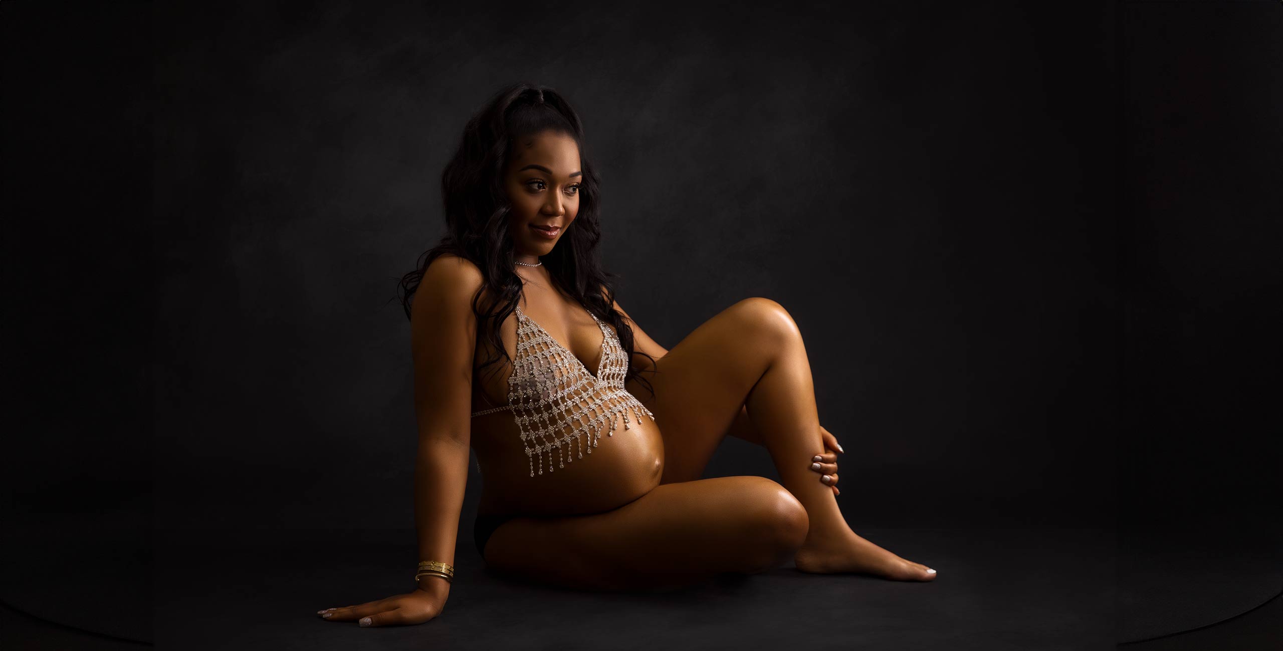 A pregnant woman with long hair sits on the floor in a studio, partaking in a maternity shoot. She is wearing a bead-adorned top and smiles softly, set against a dark background.