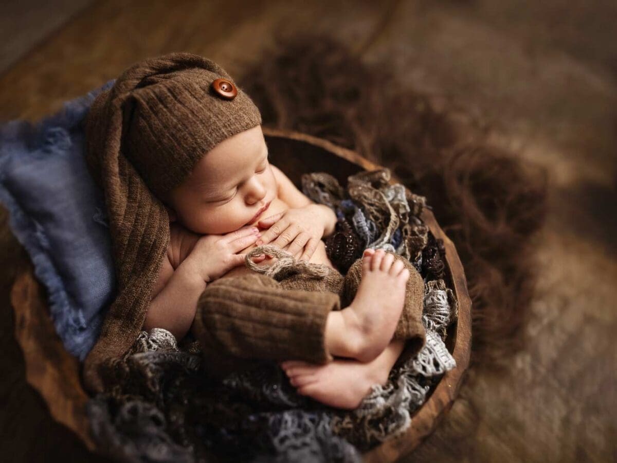 A baby sleeps in a round wooden basket, adorned in a brown knit hat and outfit, surrounded by textured fabrics—capturing the essence of newborn photography.