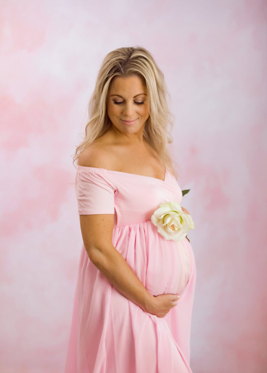 Maternity photographer Crawley studio session. Pregnant woman in a pink dress holding her belly, with a white rose tucked at her waist, set against a pink background—a perfect example of Sussex maternity portraits.