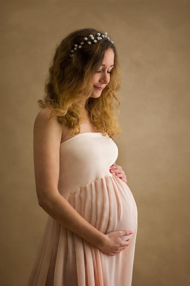 A pregnant woman with long curly hair in a cream-colored dress, gently holding her belly, captures the essence of Sussex maternity portraits as she stands gracefully against a plain background.