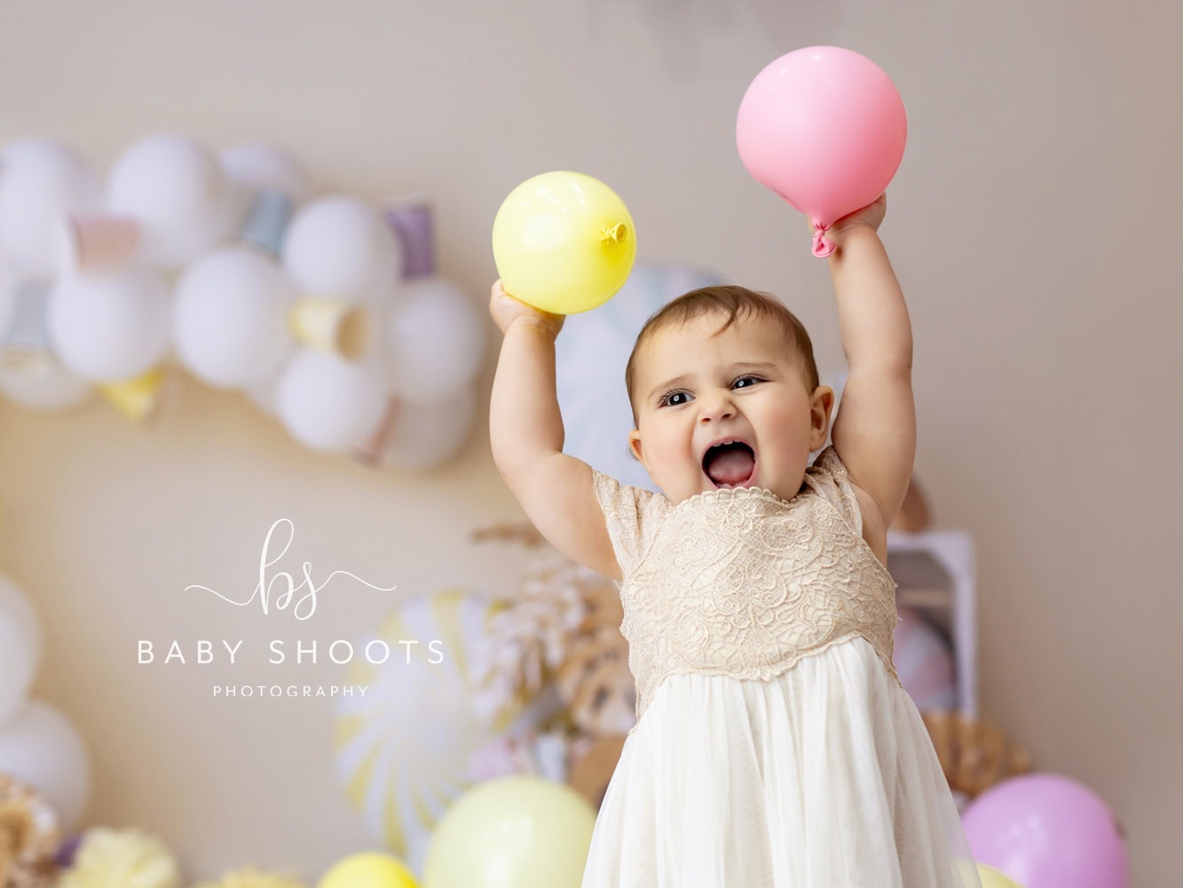 A baby wearing a cream dress holds a yellow balloon in one hand and a pink balloon in the other, surrounded by a sea of colorful balloons—perfect for capturing magical cake smash portraits in Sussex.