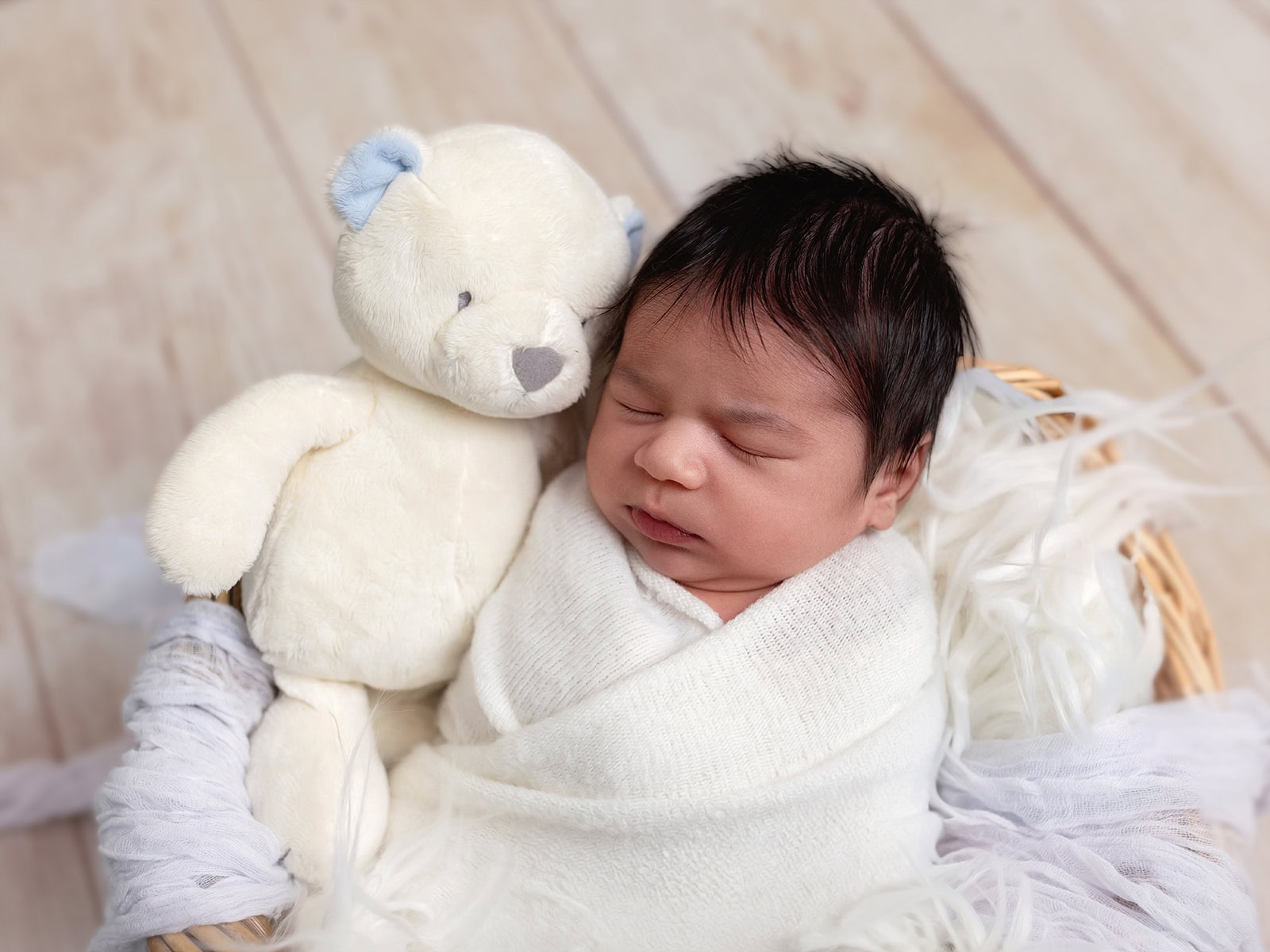 Newborn wrapped in a white blanket, sleeping in a basket with a plush teddy bear.