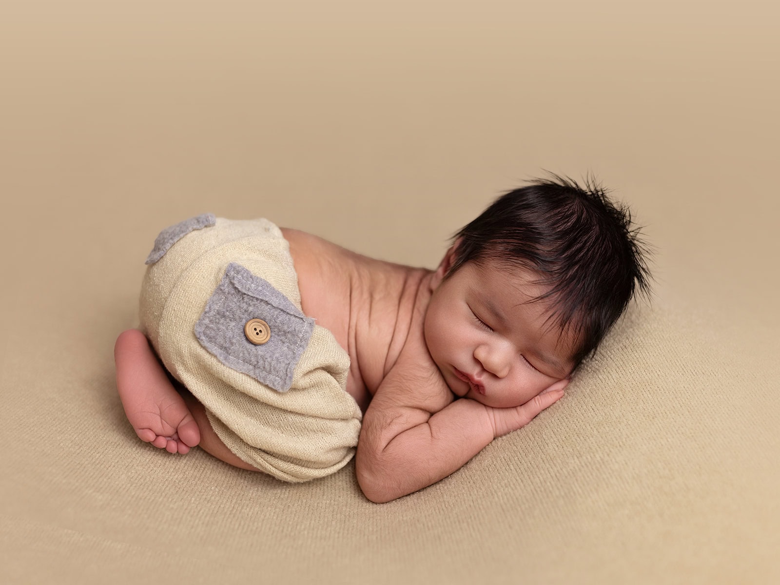 A sleeping baby lies on its side with hands under its cheek, wearing beige shorts with gray patches, on a soft beige surface.