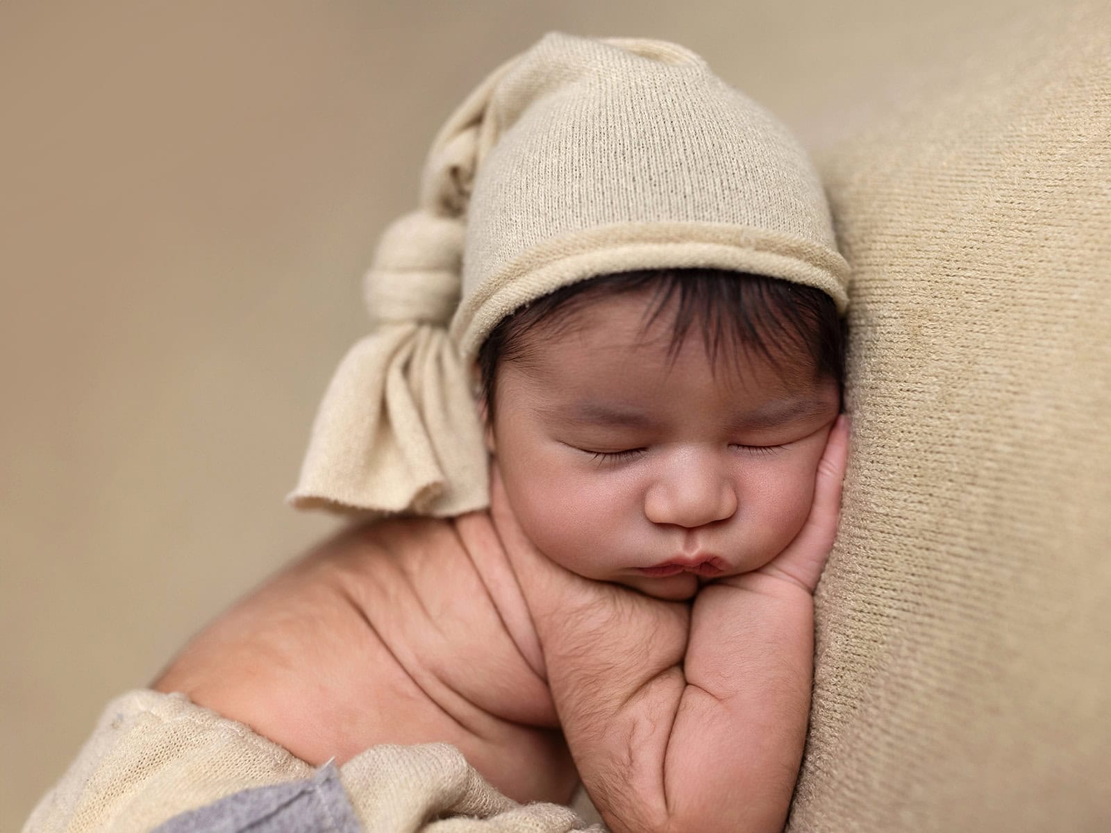 A sleeping baby rests on its side, wearing a soft beige hat with a knot. The baby’s cheeks are pressed against its hands, with a peaceful expression on its face.
