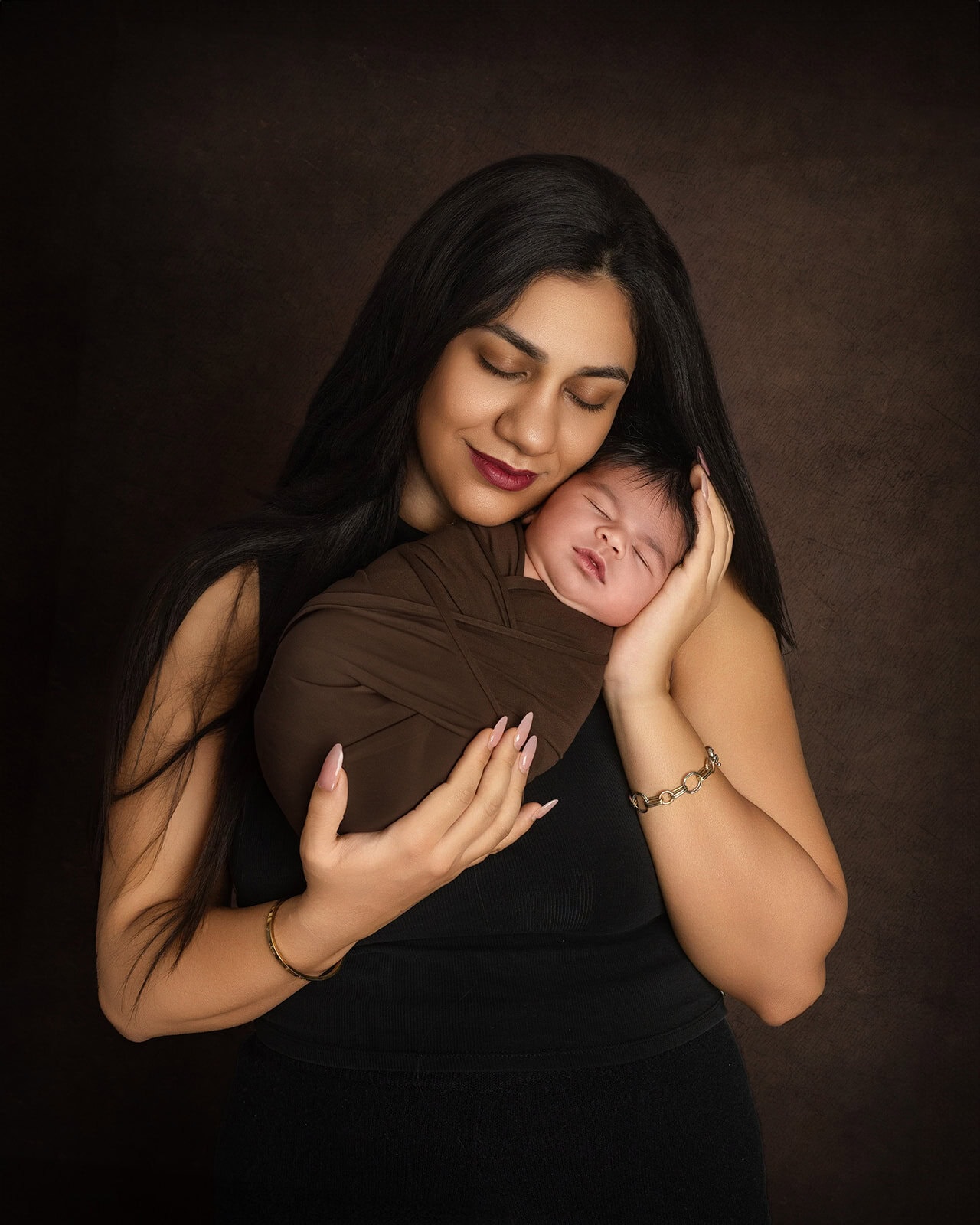 Woman with long dark hair gently holding a swaddled sleeping baby. Both have calm expressions against a dark background.