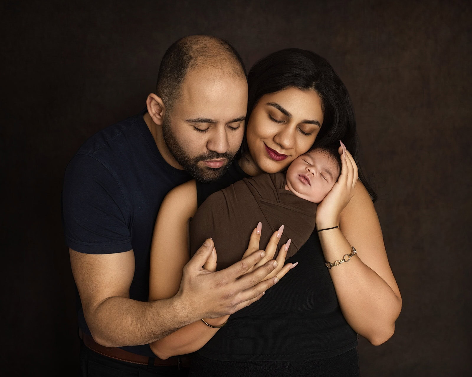 A couple lovingly holds their newborn wrapped in a brown blanket. The mother gently cradles the babys head, while the father supports the babys body.