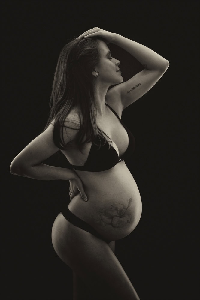 Pregnant woman in black underwear poses in profile against a dark background, one hand on her hip and the other on her head. A fetus illustration is visible on her belly.