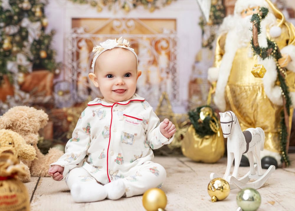 A baby in pajamas sits on a wooden floor surrounded by holiday decorations, making this the perfect setting for kids christmas portraits or an enchanting christmas mini-session. Gold ornaments, teddy bears, and a Santa Claus figure complete the festive scene.
