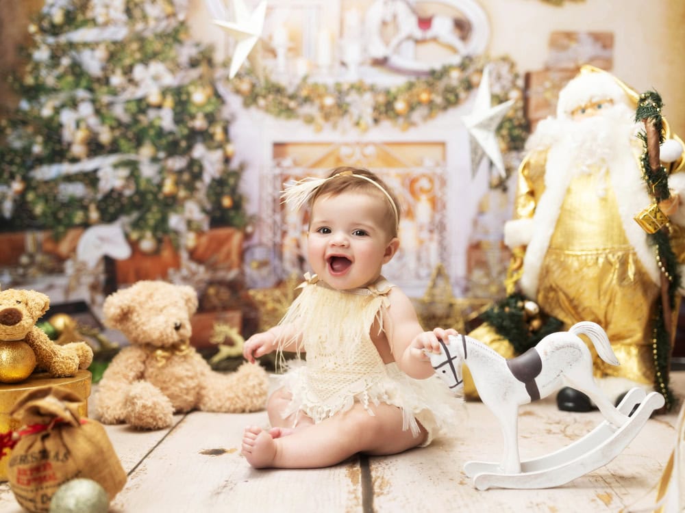 A smiling baby in a cream outfit sits on a wooden floor, surrounded by teddy bears, a toy rocking horse, and festive decor—perfect for kids Christmas portraits during your christmas mini-session. A Christmas tree and Santa figure add holiday cheer in the background.