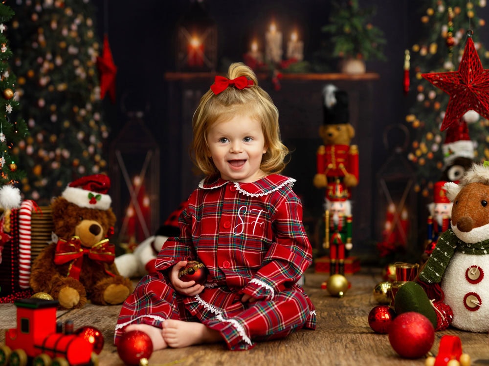 A young child in red plaid pajamas sits on a wooden floor with Christmas decorations, stuffed animals, and a toy train—perfect for capturing magical kids Christmas portraits in front of a festive holiday backdrop.