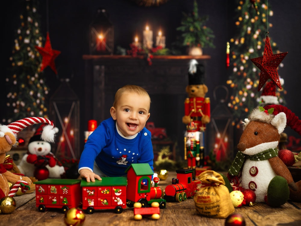 A smiling baby in a blue sweater plays with a toy train, surrounded by festive decorations—perfect for kids Christmas portraits or christmas mini sessions by the fireplace and sparkling Christmas trees.
