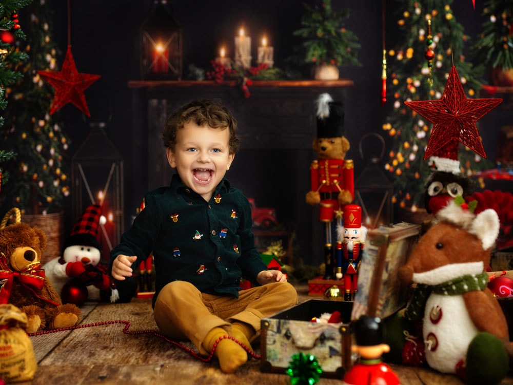 A young boy sits smiling on a wooden floor surrounded by Christmas decorations and toys, perfect for kids Christmas portraits, with a decorated tree, glowing fireplace, and candles in the background.