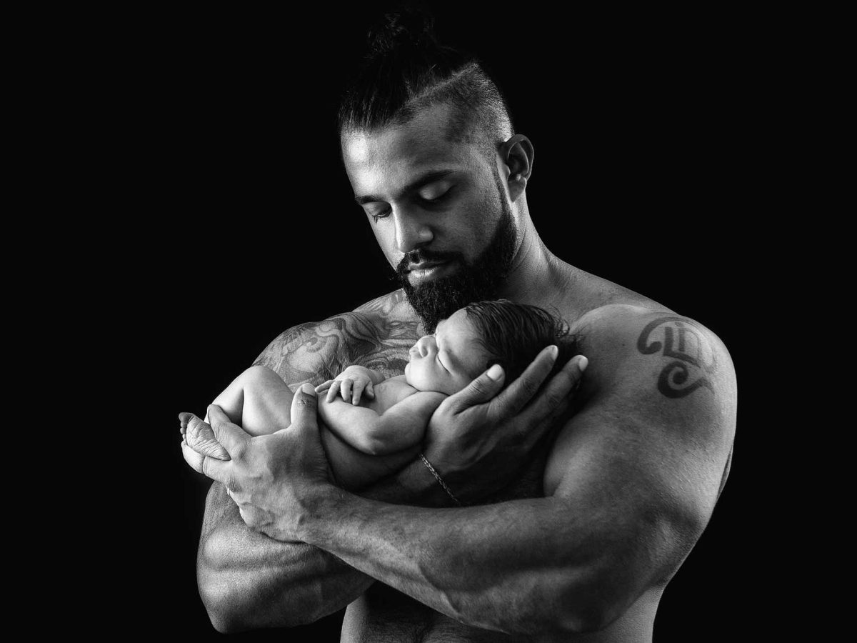 A stylised newborn photoshoot featuring a muscular father holding his small baby, with creative directional lighting accentuating form, contrast, and the scale between them.
