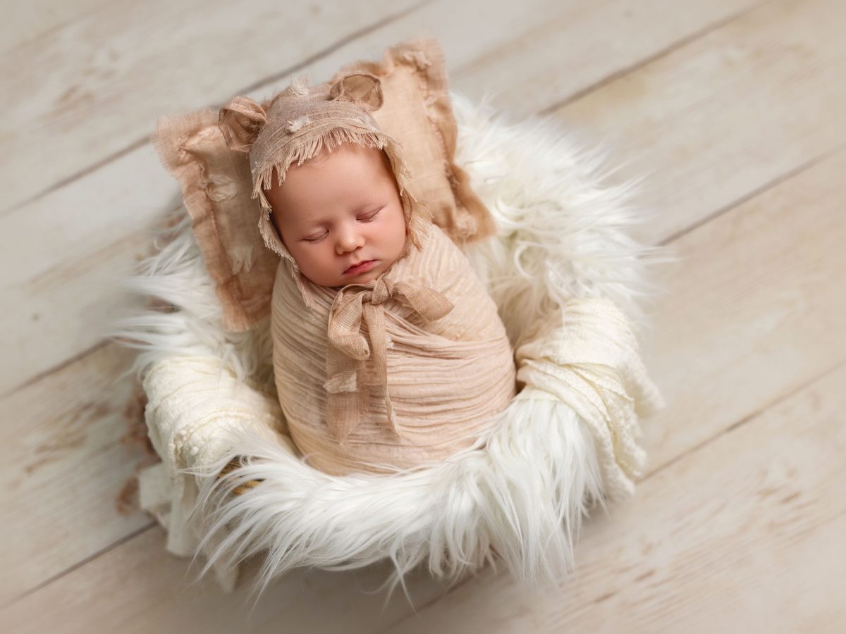 Fine art baby portraits for families in Merstham RH1 featuring a fully wrapped newborn baby sleeping in a soft and furry basket prop with bonnet and a light-coloured wooden floor.