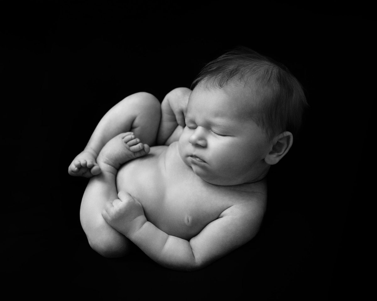 Fine art newborn photography Nutfield and Bletchingley featuring a b/w newborn poirtrait of baby sleeping in a natural, curly pose, backlit against a black background.