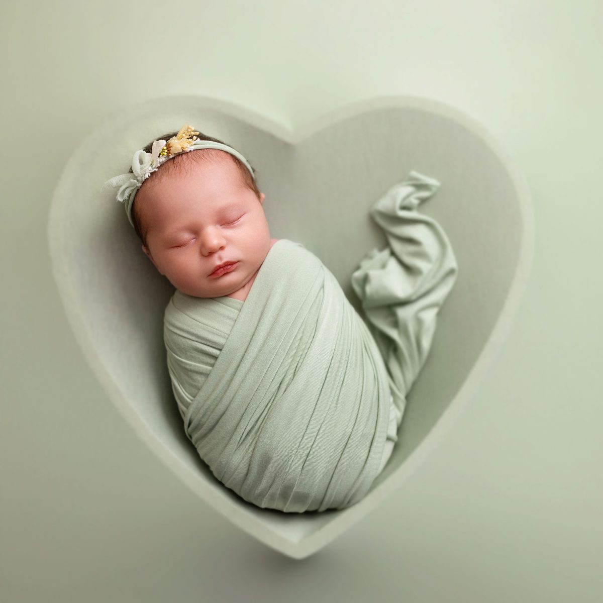 Creative newborn art at our studio near Horley, featuring a baby posed with delicate floral headband sleeping in a heart-bowl with matching green wrap.