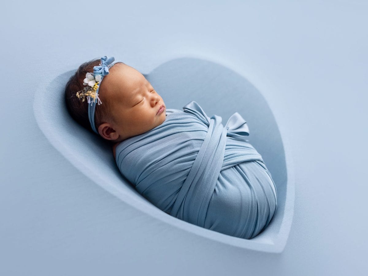 Creative newborn portraits for parents in South Godstone, featuring a beautiful portrait of a sleeping baby snuggly wrapped in blue with a matching floral headband, and laying in a baby-blue heart-shaped bowl.