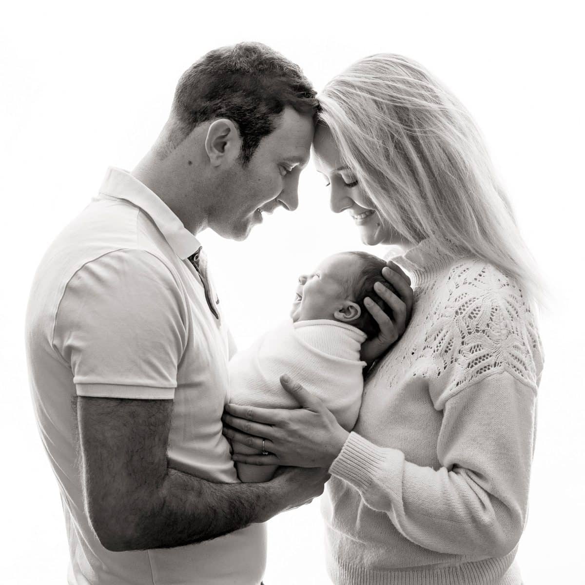 A high-key family portrait taken during a newborn photoshoot in Sussex. This black and white image is backlit for a soft, and dreamy effect, for this beautiful family newborn portrait with both parents and baby.
