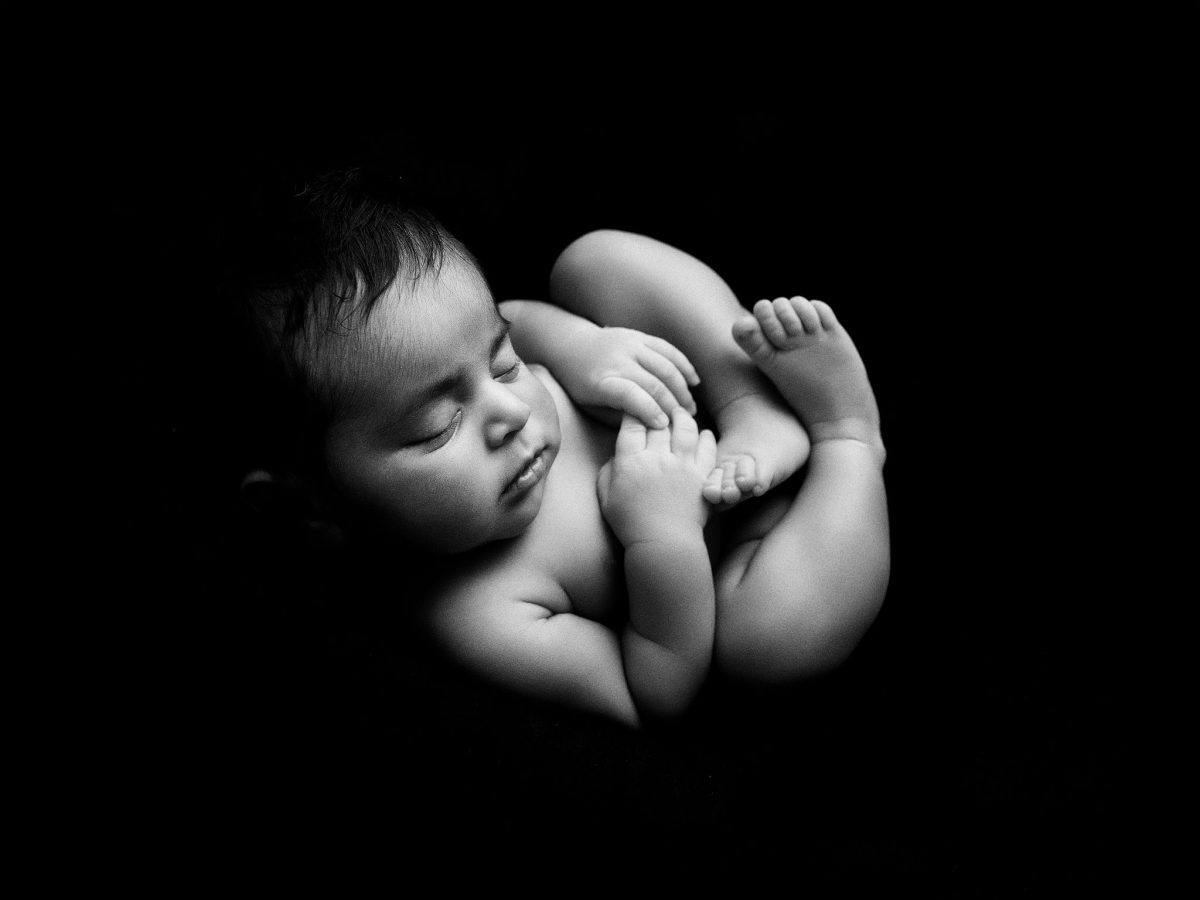 Fine art baby photographers for parents in Horley, featuring a newborn in a comfortable curly pose on a black background in a b/w fine-art style.