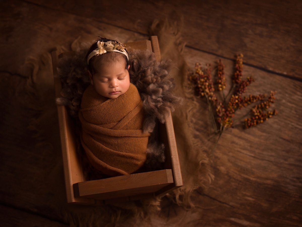 Fine art newborn portraits for families in Meath Green, Horley, featuring a creative low-key portrait of a baby laying in a rustic wooden prop with warm brown tones and textures.