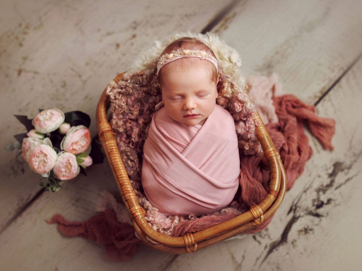 Luxury newborn portraits for families in the Folders Lane area of Burgess Hill, featuring a sleeping baby with a floral headpiece asnd matching flowers next to a quaint wicker basket.