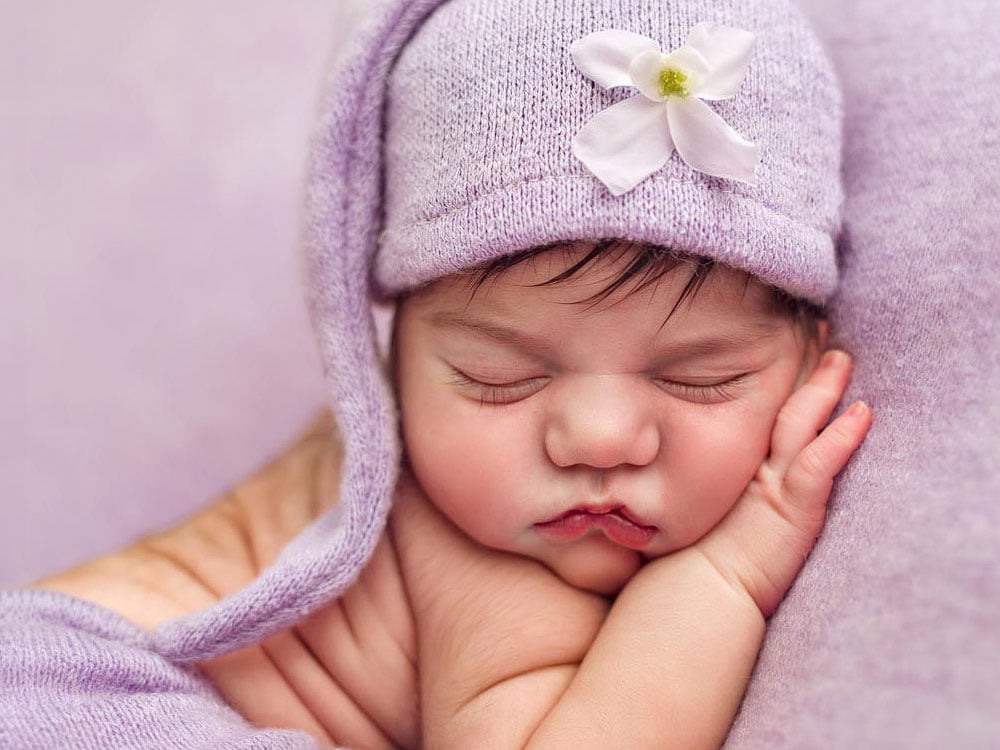 Natural newborn photography in Copthorne, Crawley. A newborn peacefully sleeps, dressed in a purple outfit adorned with a flowered hat, resting snugly on a matching purple blanket. Newborn portraits by Sussex newborn photographers Baby Shoots Photography