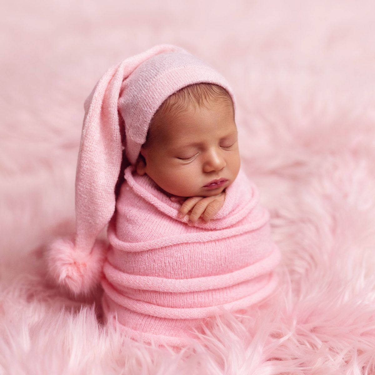 Boutique newborn photography packages for Horsham (RH12) families: A baby sleeping soundly, in a soft pink wrap with matching hat, on a soft, furry rug.