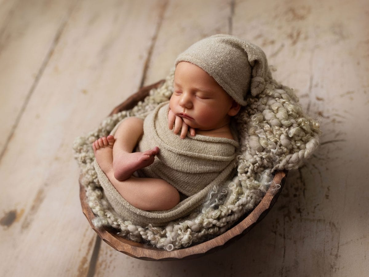 High-end newborn photography packages in the East Grinstead area: A baby sleeping soundly in a wooden bowl with matching wrap and hat on a rustic wooden floor.