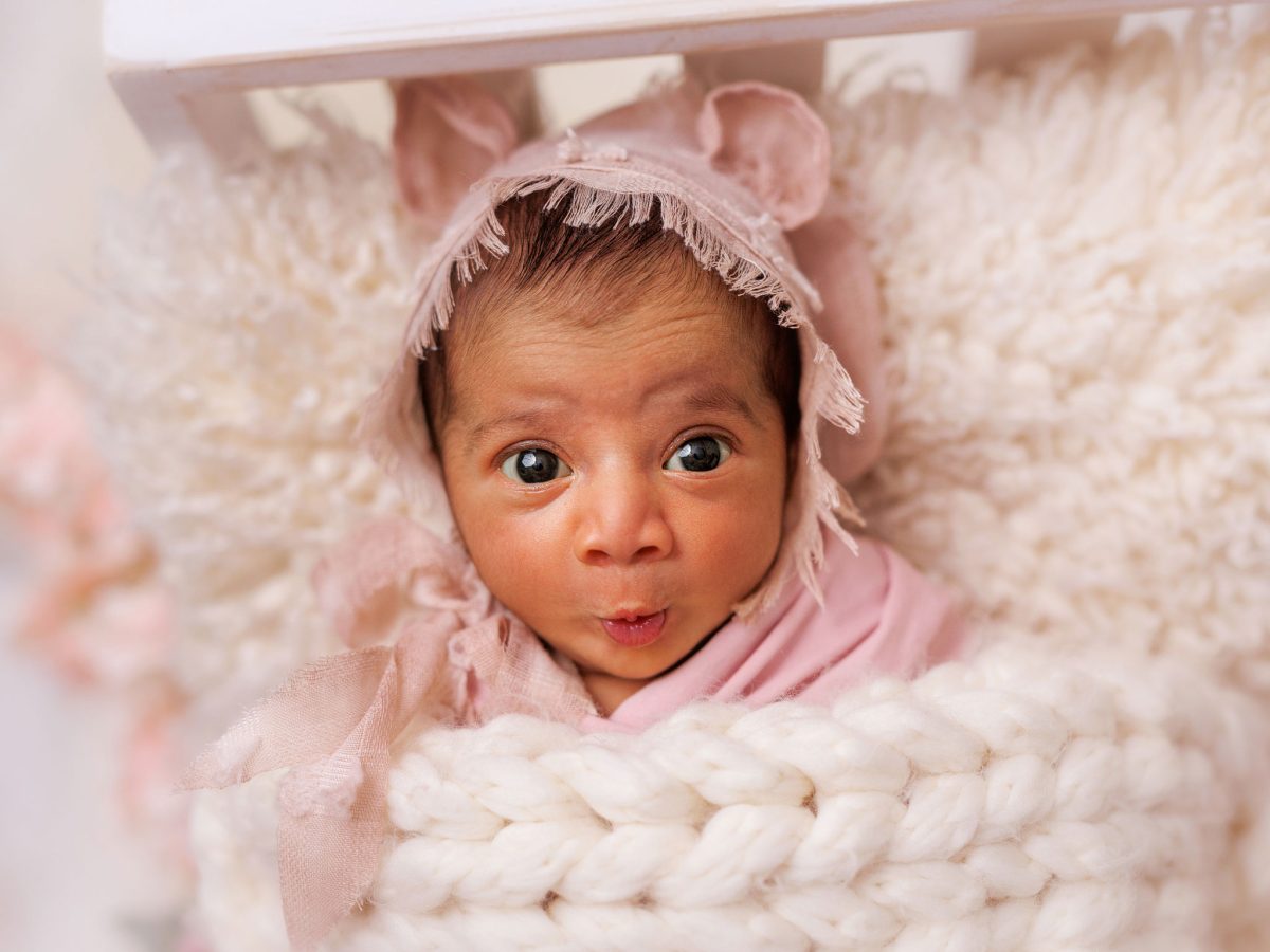 Safety-centred newborn photographer serving Haywards Heath, showing a baby comfortably posed in a bespoke bed prop, with a comical expression on her very cute face..