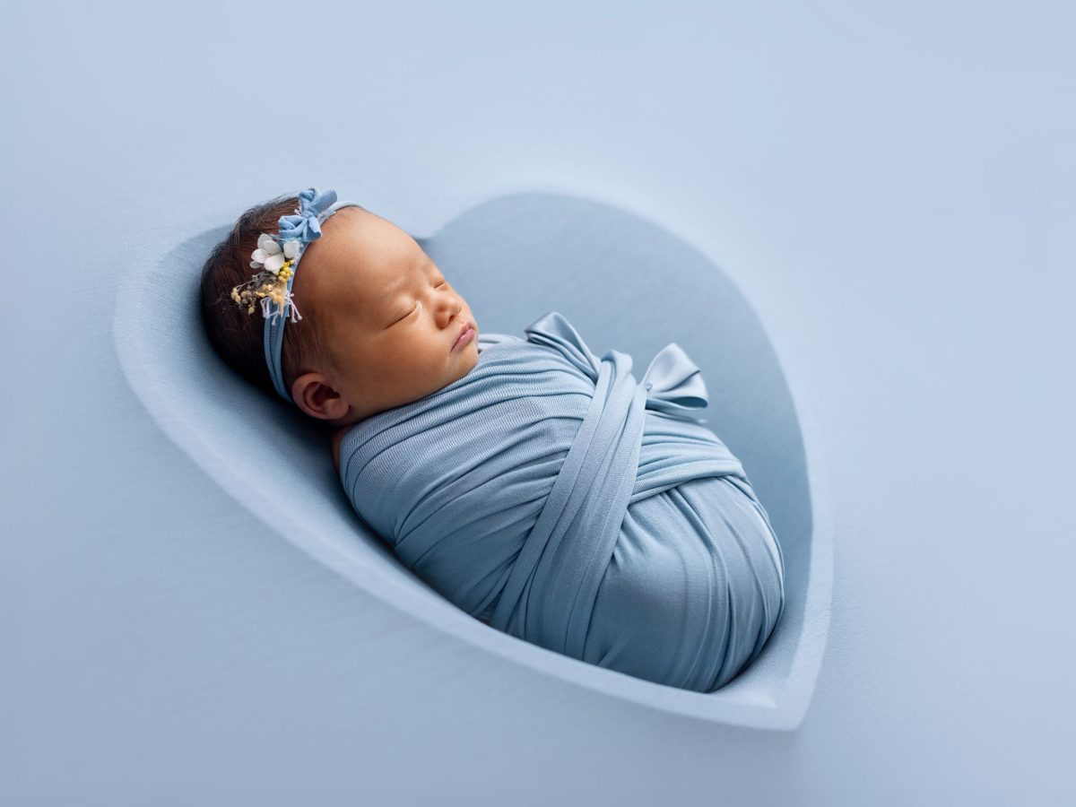 A simple, backlit portrait of a newborn baby, wrapped in a soft blue wrap on a blanket outlined by a heart-shape