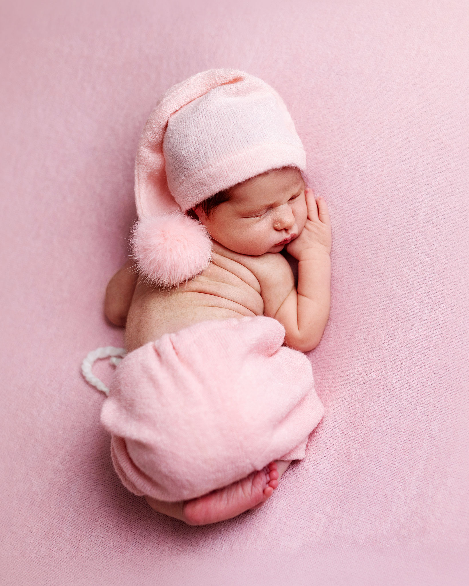 Sleeping newborn photographed with soft natural-style lighting on a pink blanket in studio.